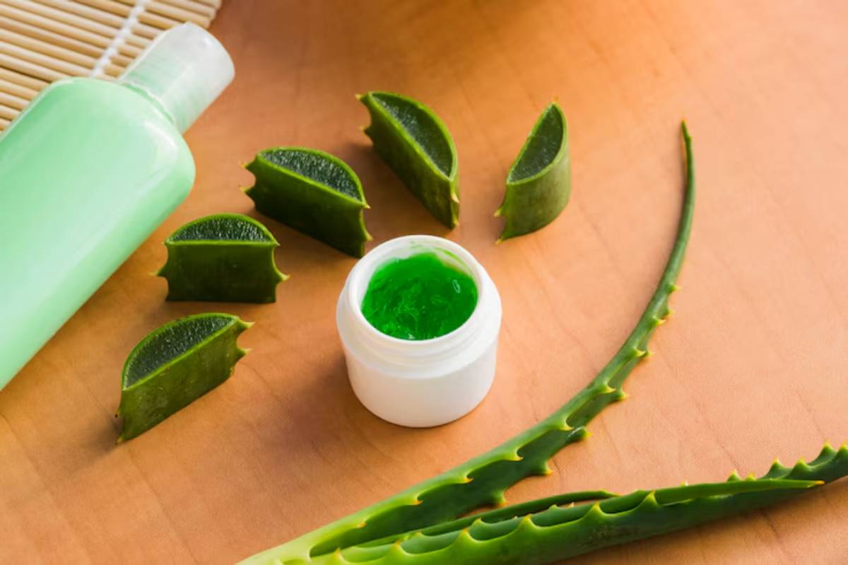 Aloe vera gel in a white jar surrounded by sliced aloe leaves and a green bottle on a wooden surface, showcasing natural skincare.