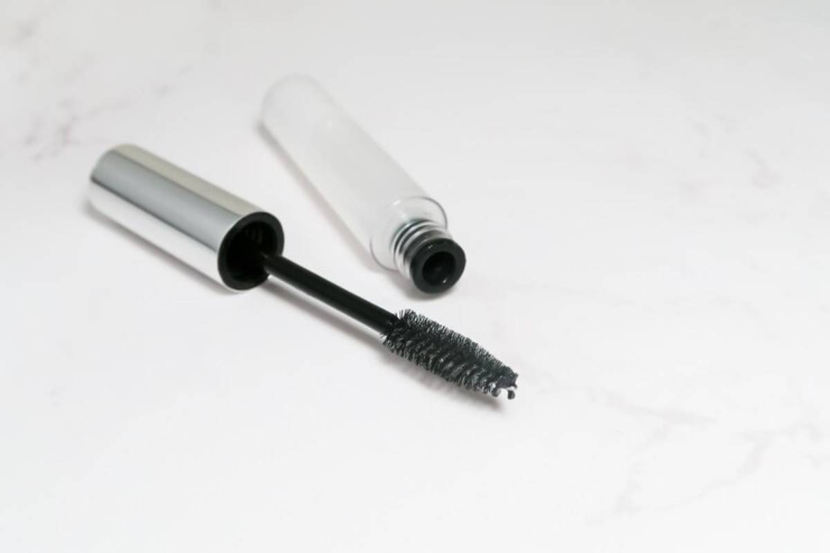 An open mascara tube with a black wand, resting on a light marble surface. The sleek silver cap contrasts with the clear body.