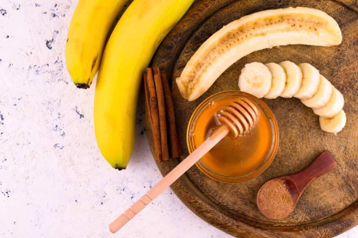 A wooden tray with bananas, honey in a jar, sliced bananas, a cinnamon stick, and a spoon with cinnamon powder.