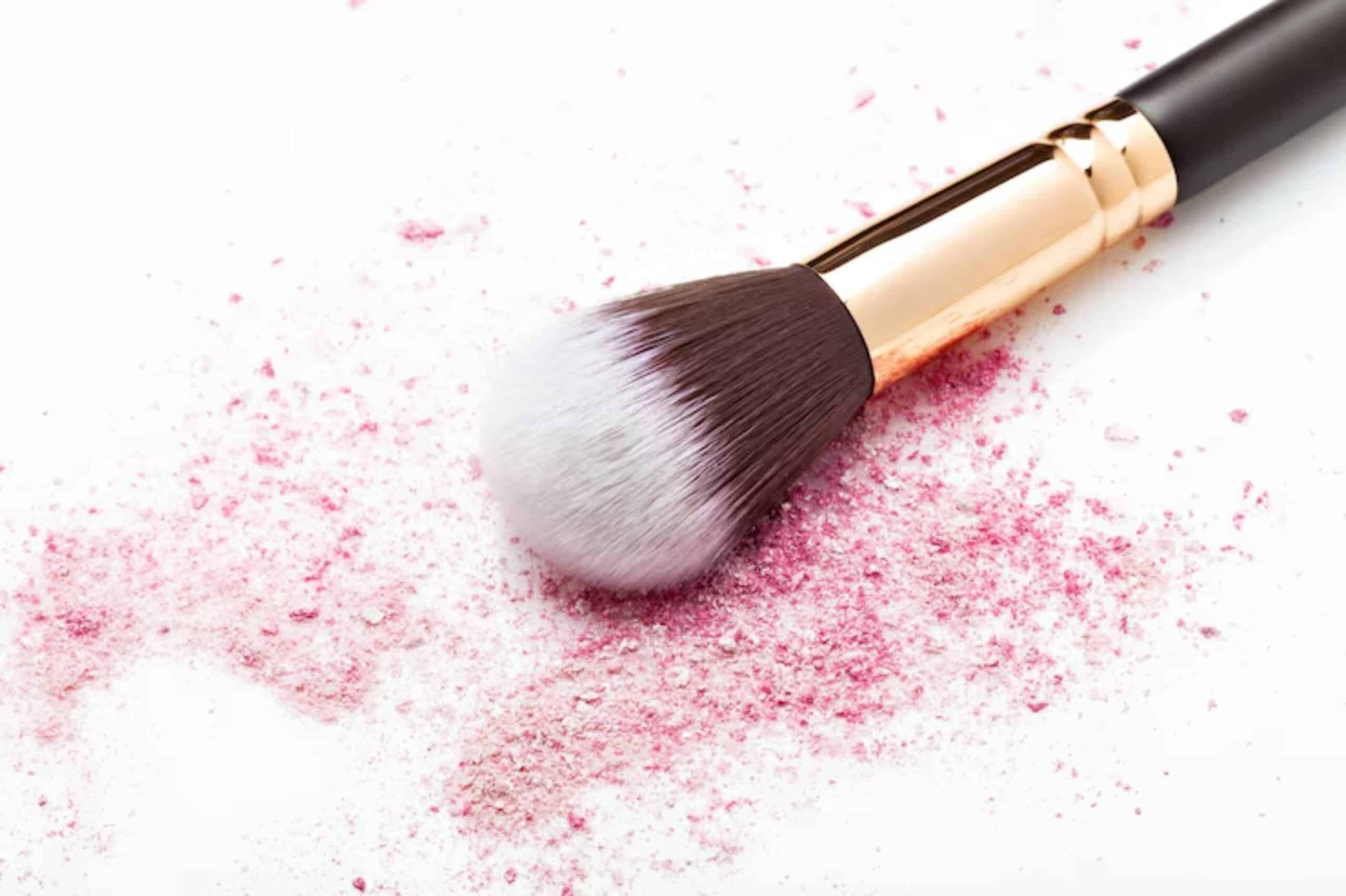 A makeup brush with a pink tip rests on a surface scattered with pink powder, creating an artistic and messy beauty scene.