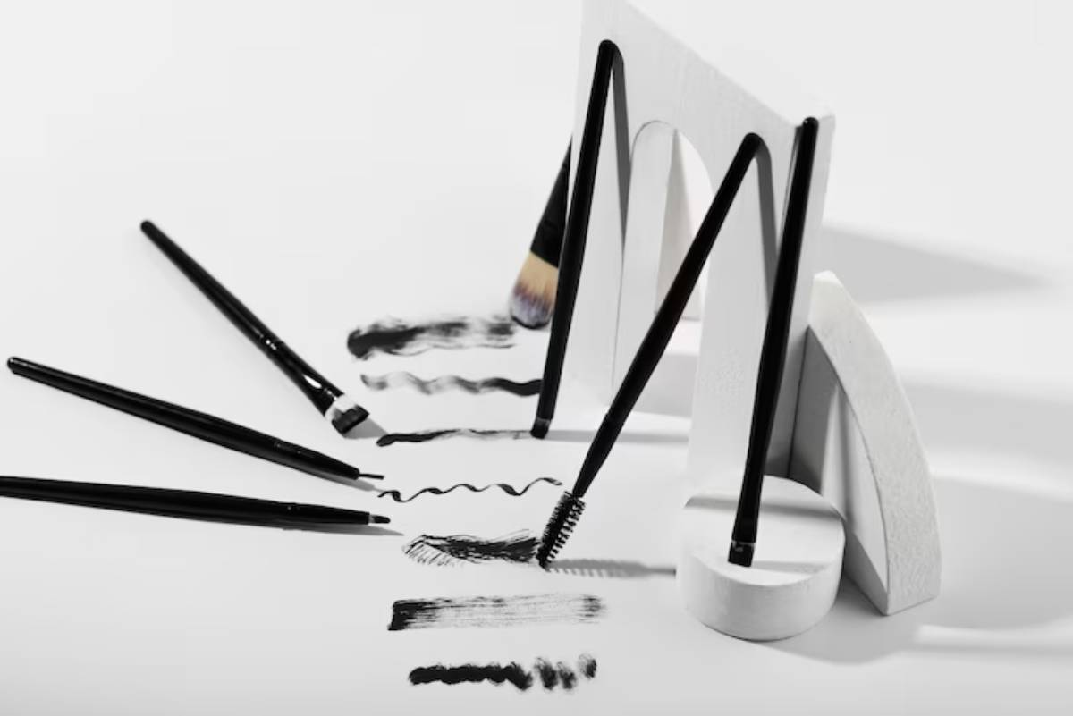 Artistic arrangement of makeup brushes and pencils creating black strokes on white paper, with geometric props in the background.