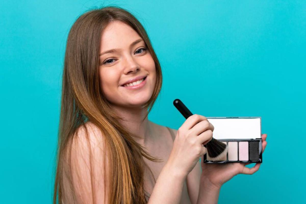 A person holds a makeup palette with a brush against a vibrant turquoise background, preparing to apply makeup.