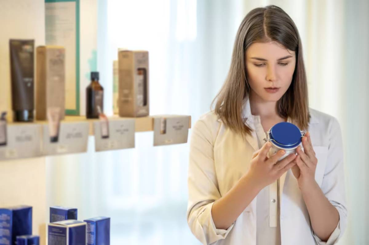 A person in a white coat holds a jar with a blue lid, surrounded by various beauty and skincare products on display.