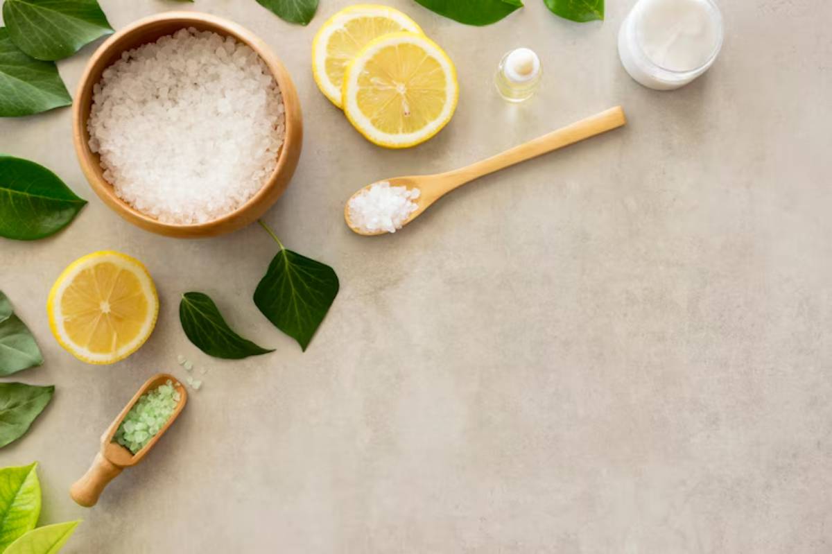 A wooden bowl filled with salt, sliced lemons, leaves, a small scoop of green salt, and a jar of cream on a gray background.