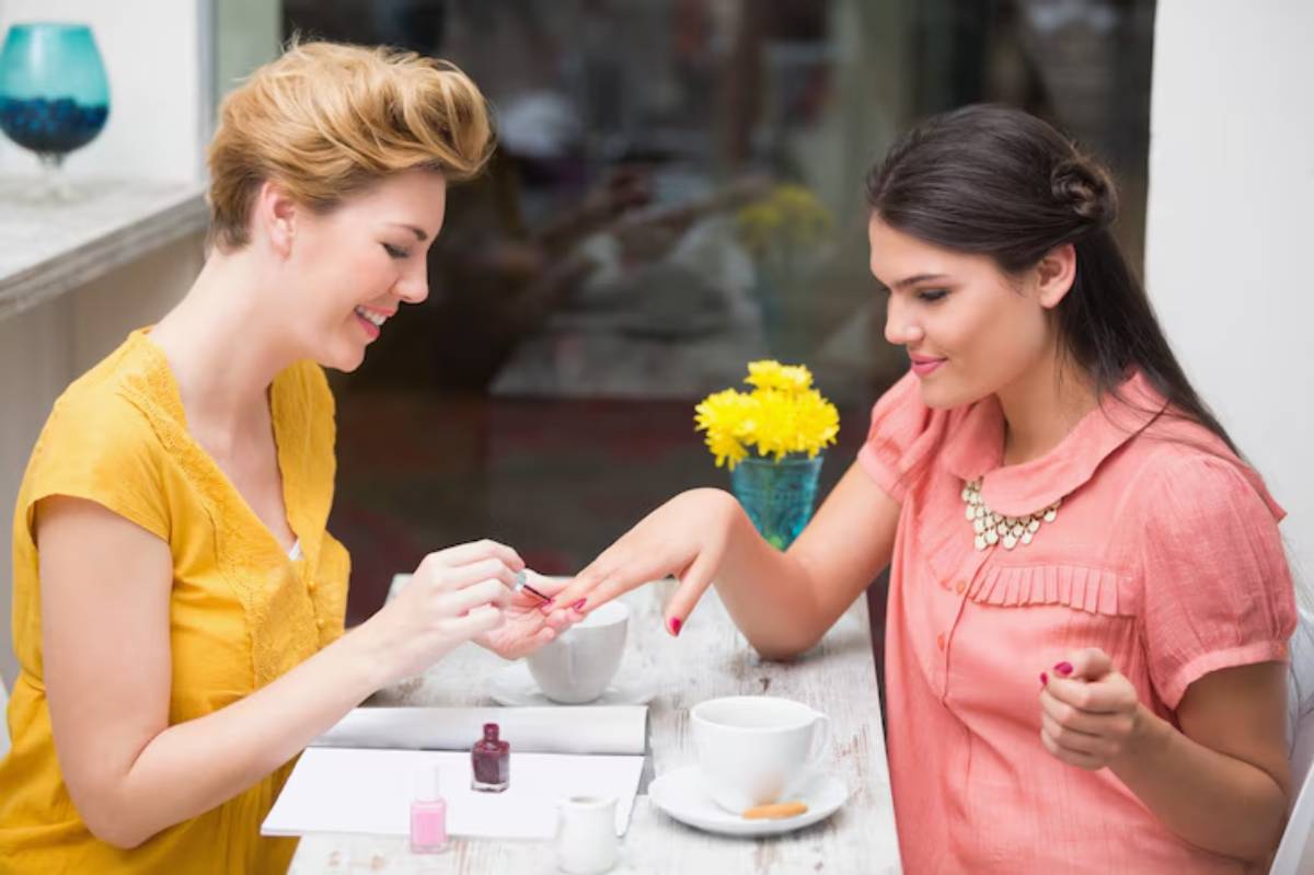 A woman in a yellow shirt applies nail polish to another woman's hands, surrounded by flowers and coffee cups in a bright setting.