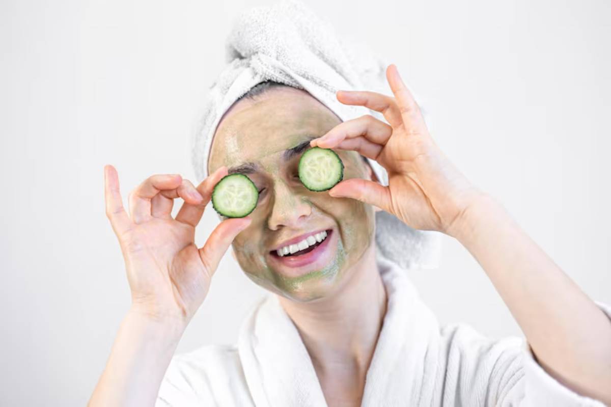 A person wearing a white robe and towel on their head holds their hands up as if applying a product, set against a plain background.