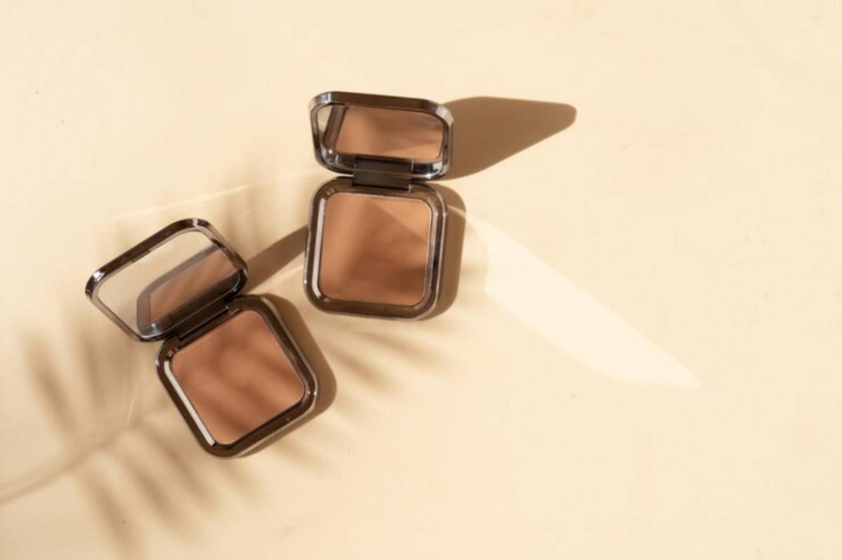 Two open compact bronzers sit on a light beige surface, casting soft shadows, with a hint of palm leaf shadow in the background.