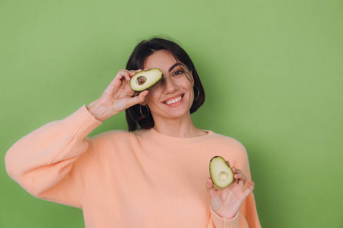 A person in a pastel pink sweater holds a halved avocado against a vibrant green backdrop.