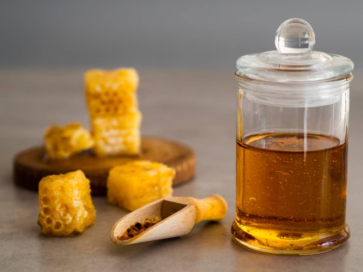 A glass jar of honey next to pieces of honeycomb on a wooden board and a wooden scoop filled with bee pollen, all against a neutral background.