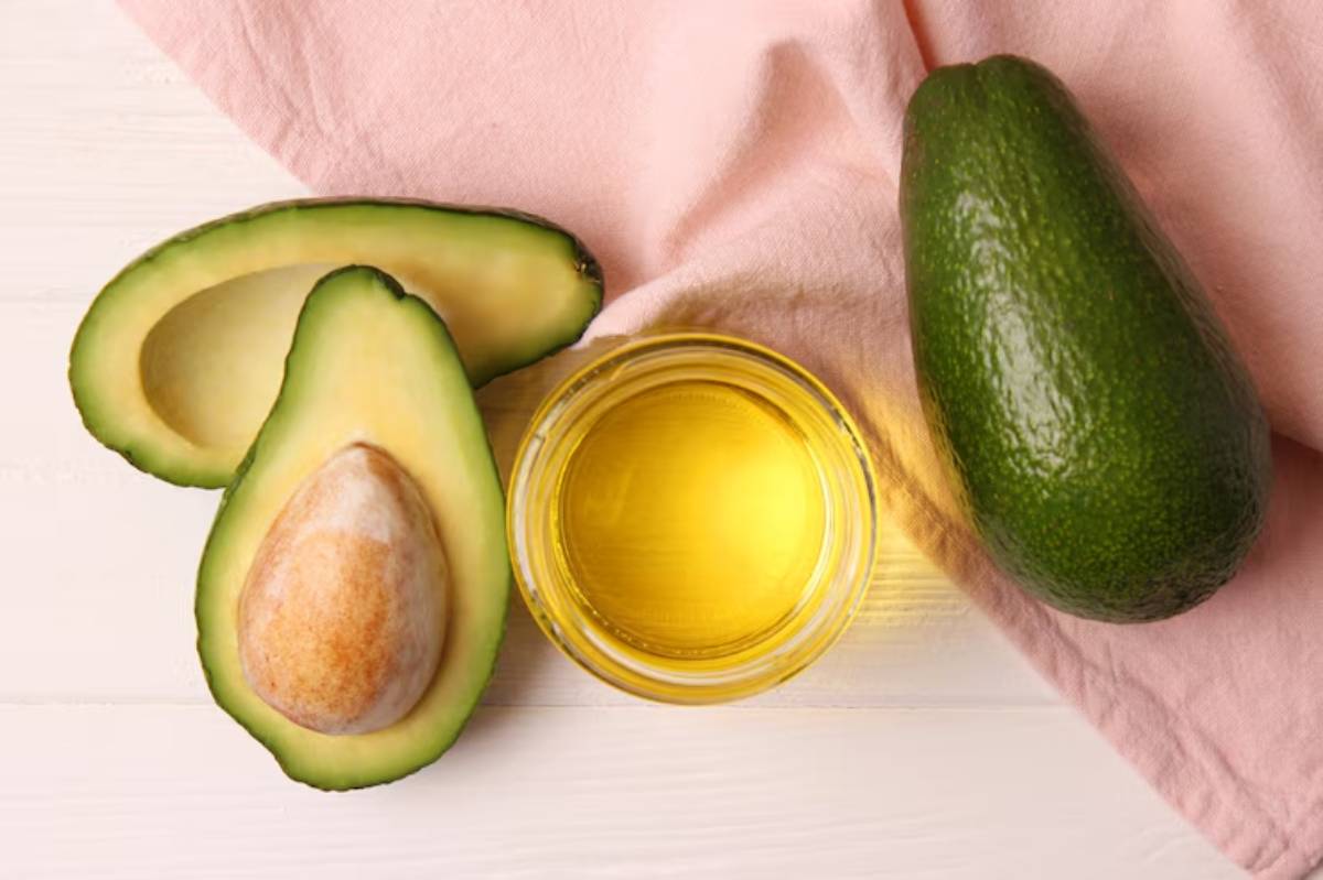 Fresh avocado halves with a seed, whole avocado, and a jar of golden avocado oil on a pink textured background.