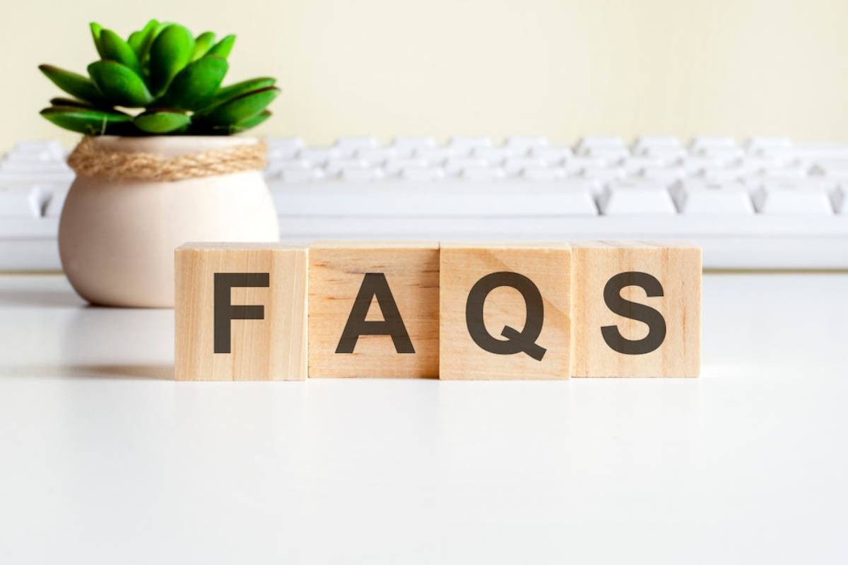 Wooden blocks spelling FAQs next to a small potted plant, with a blurred keyboard in the background.