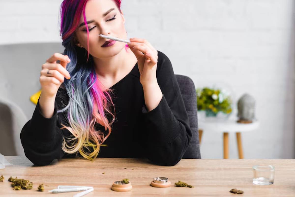 A person with vibrant, multi-colored hair sits at a table with herbs, rolling papers, and a small glass container, engaged in preparation.