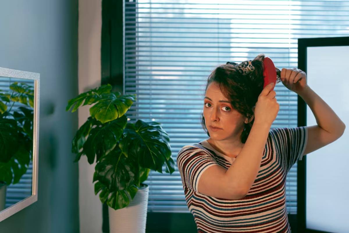 A person wearing a striped shirt brushes their hair in front of a mirror, with a green leafy plant nearby and sunlight streaming through blinds.