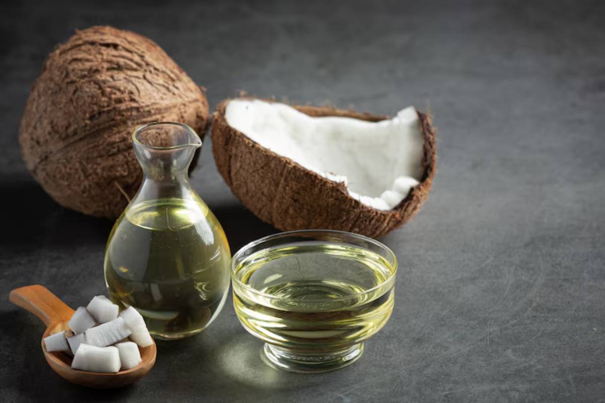 A glass jug of coconut oil, a bowl of liquid coconut oil, and pieces of coconut are arranged with a split coconut shell on a dark surface.