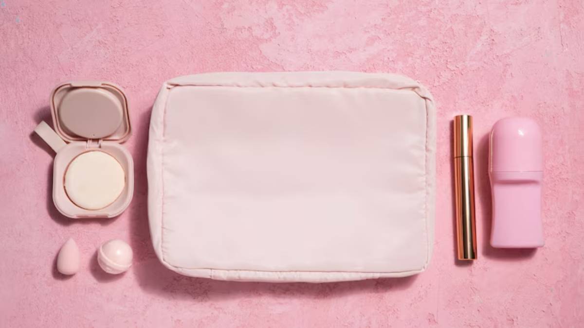 Flat lay of a light pink cosmetic pouch, compact, mascara, deodorant, and beauty sponges on a textured pink background.