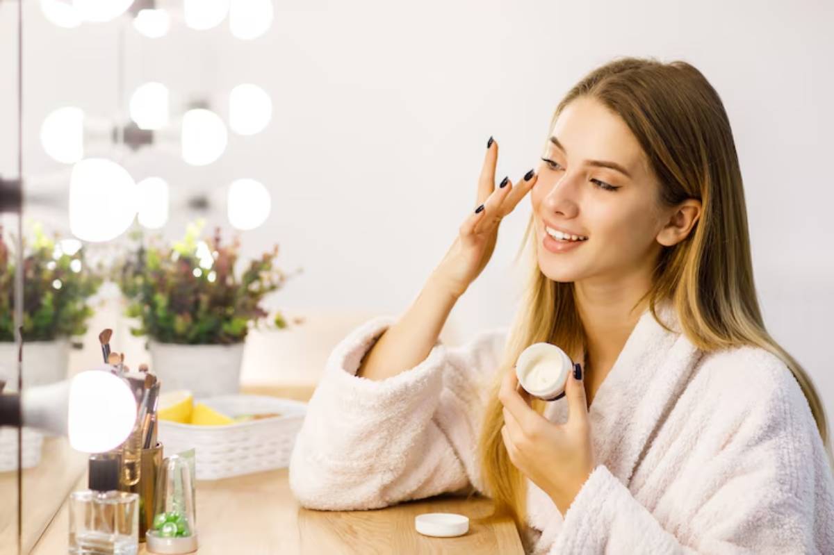 A woman in a cozy robe applies cream while seated at a vanity, illuminated by soft lighting, with plants and beauty products in view.