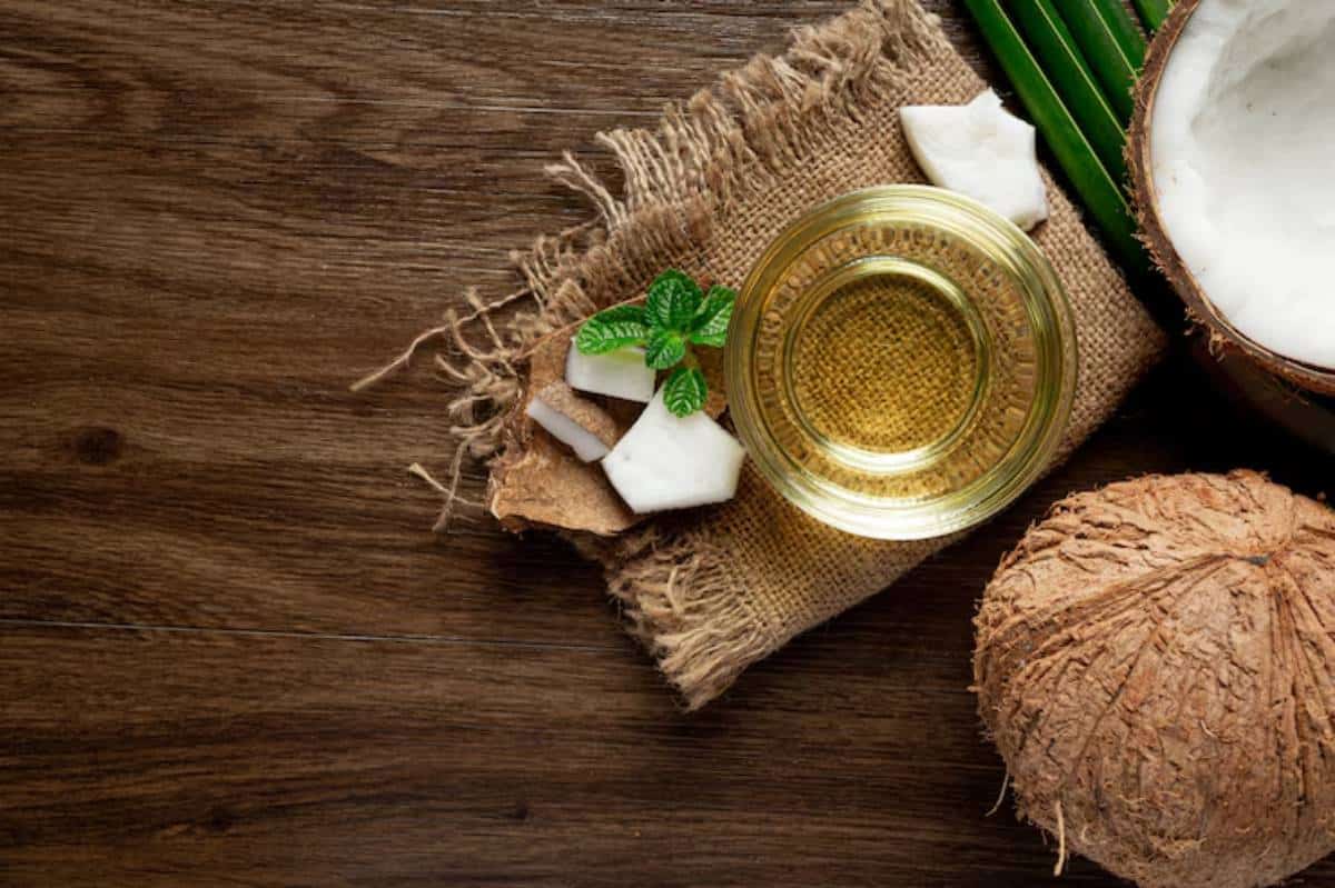 A glass jar of coconut oil, fresh coconut pieces, and green leaves on a burlap cloth, set against a wooden background.