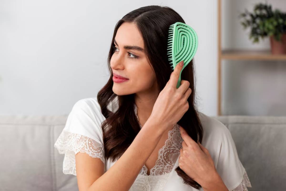 A woman with long, wavy hair gently brushes her hair with a green comb while sitting on a light-colored couch.