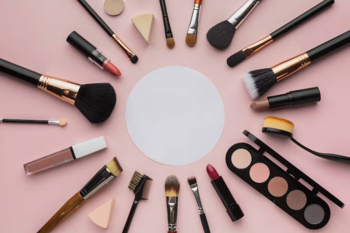 An assortment of makeup brushes, lipsticks, and a cosmetic palette arranged around a blank circular space on a pink background.