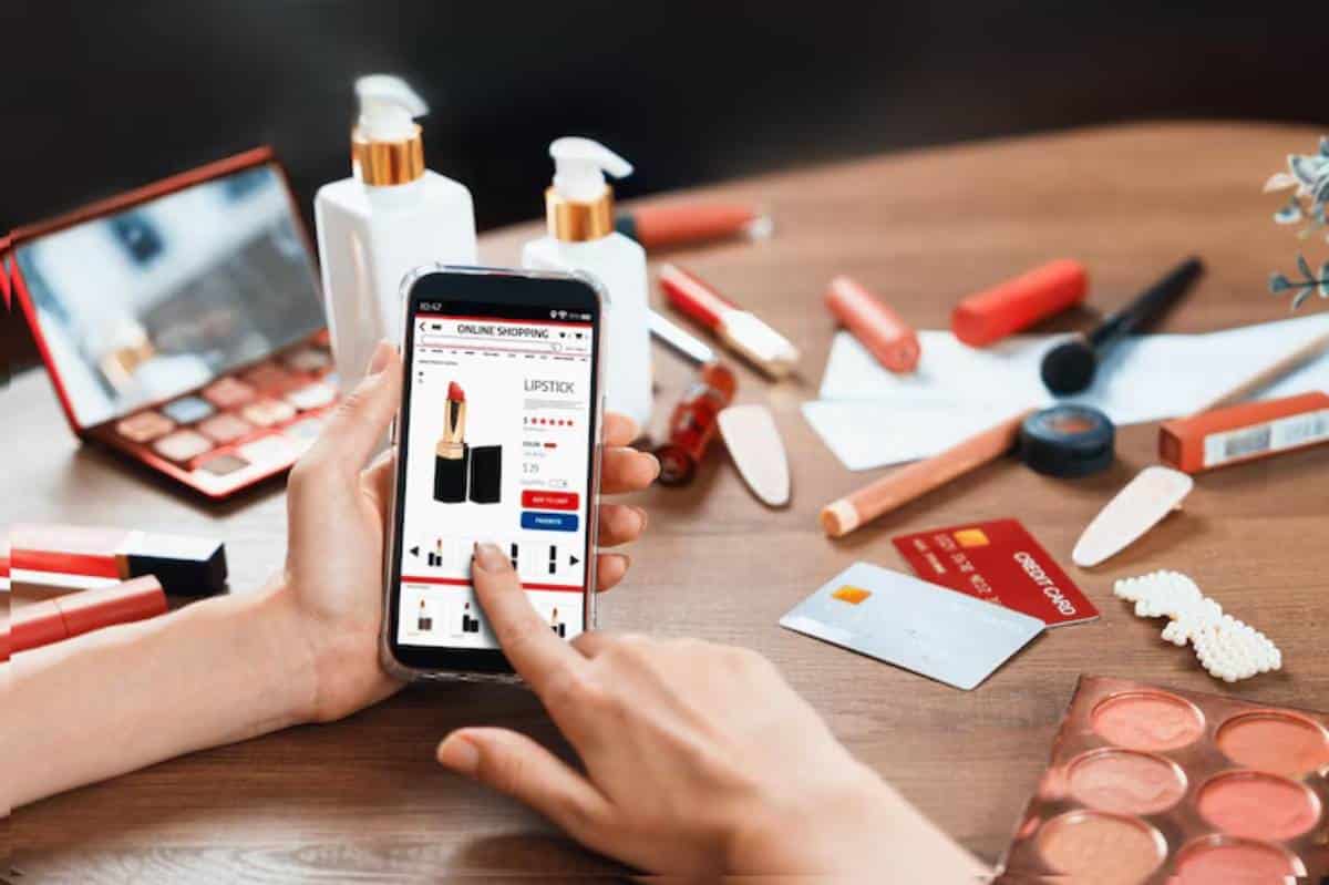 A person scrolling through an online makeup store on a smartphone, surrounded by various cosmetics and credit cards on a wooden table.