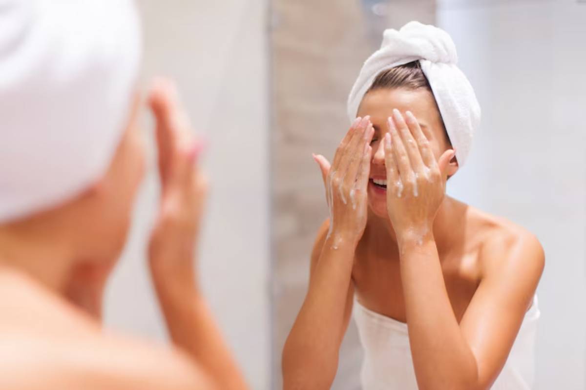 A woman with a towel wrapped around her hair gently washes her face in a bright, modern bathroom.