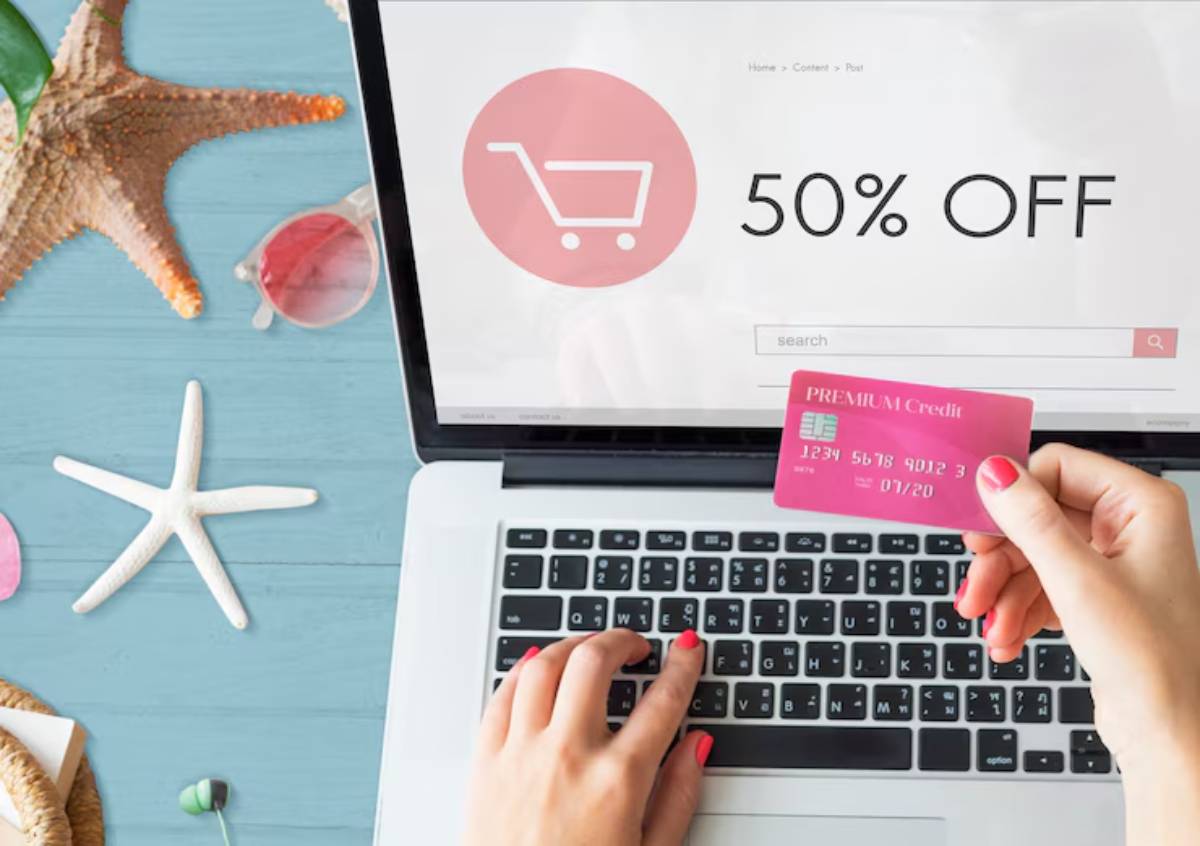 A person holding a credit card while shopping online, with a 50% OFF banner visible on the laptop screen. Beach-themed decor nearby.