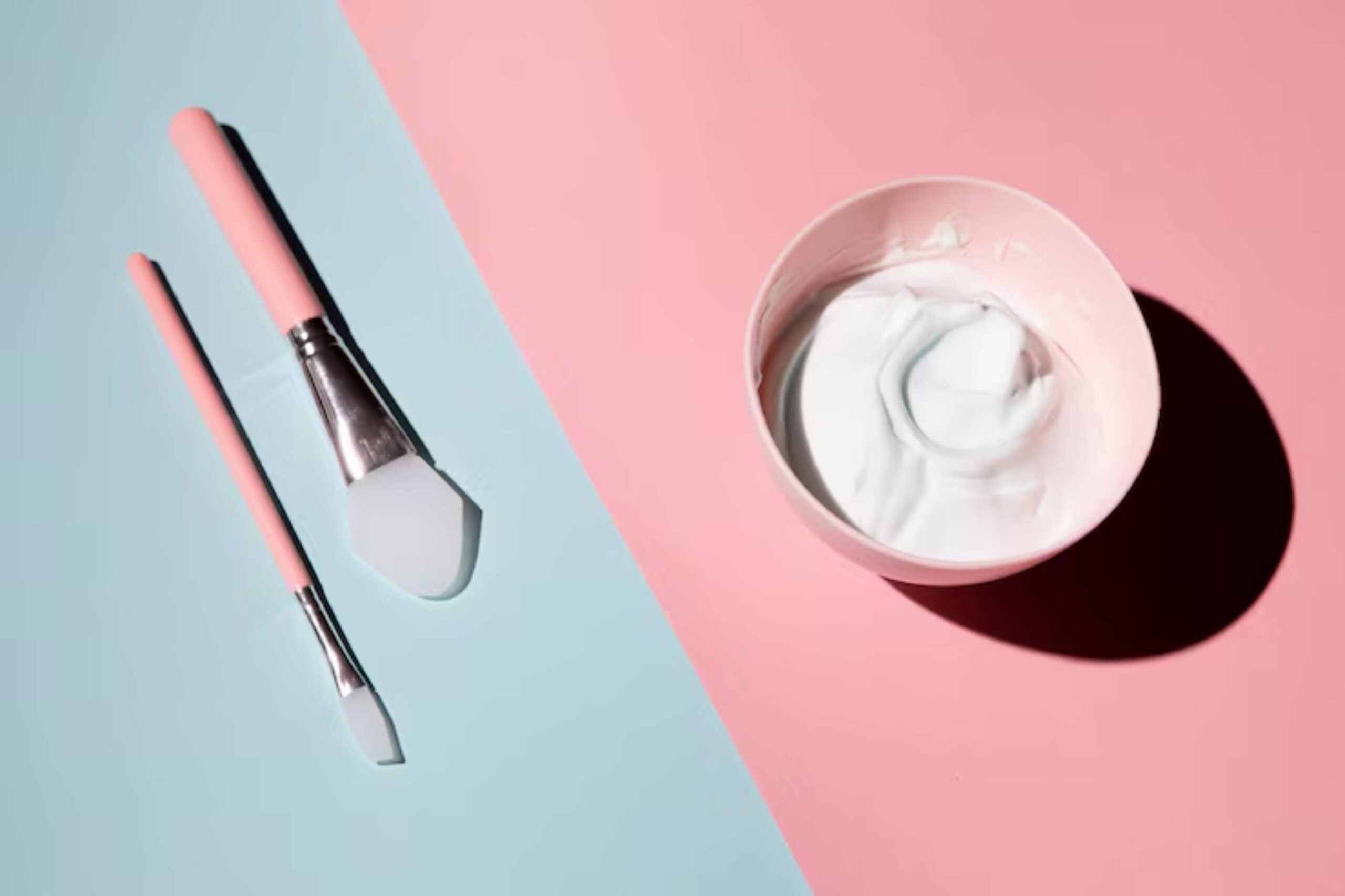 Two pink brushes and a bowl of white cream on a pastel blue and pink background, suggesting a skincare or beauty setting.