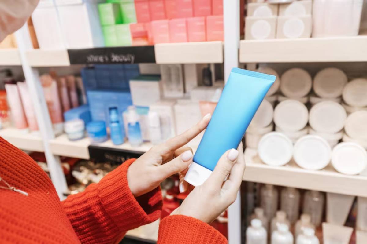 A person in a red sweater examines a blue skincare tube in a well-stocked beauty aisle filled with various products.