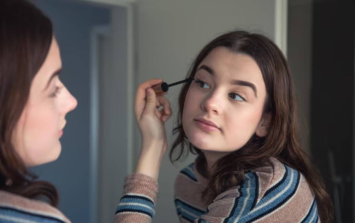 A person applies mascara while looking in a mirror, showcasing a cozy indoor setting with neutral wall colors.