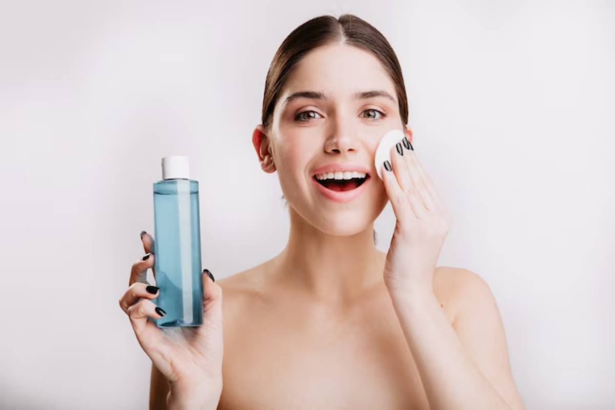 A woman holds a blue bottle of liquid in one hand while gently touching her face with the other, set against a white background.
