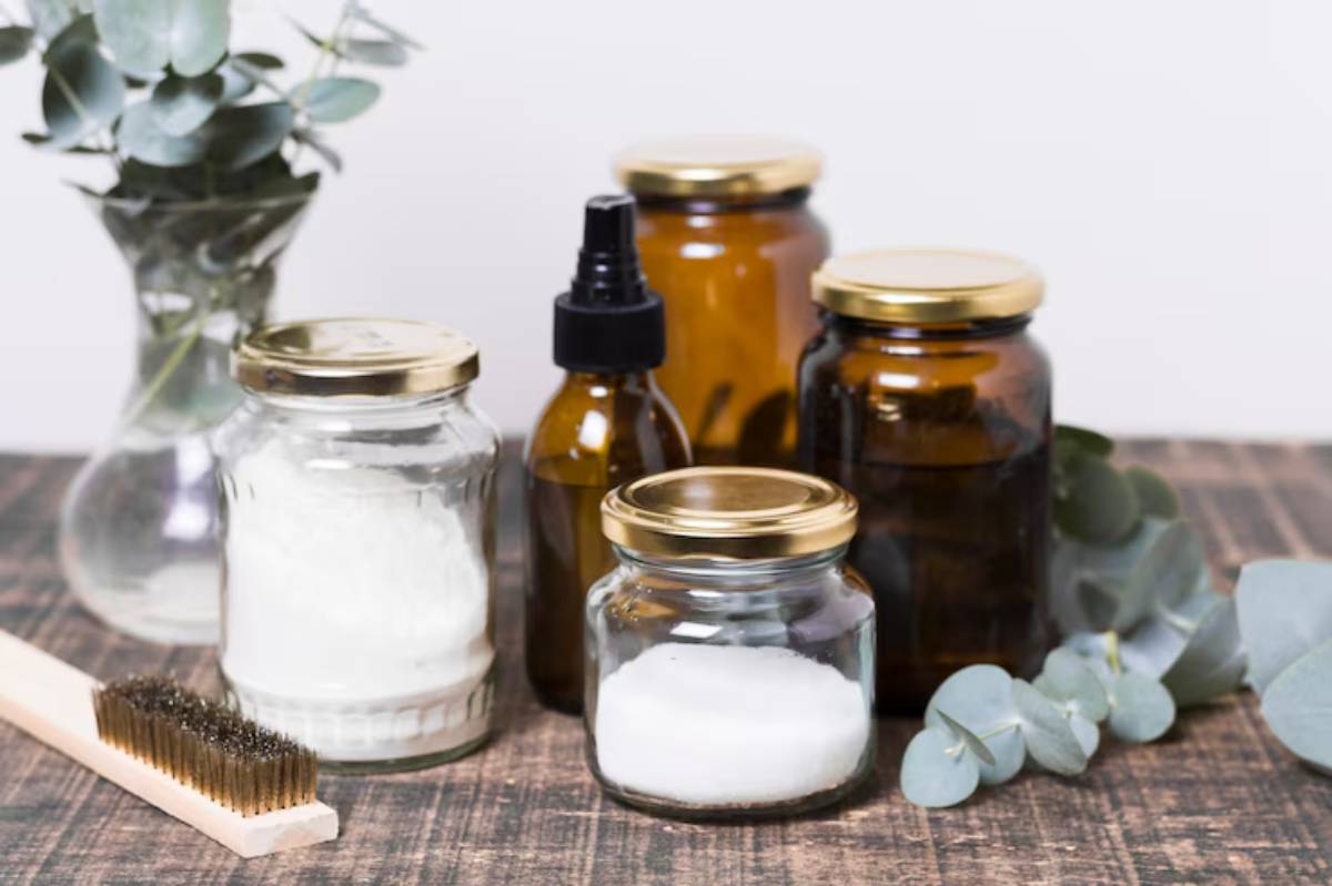 A collection of glass jars with gold lids, containing various ingredients, alongside a wooden brush and fresh eucalyptus leaves.