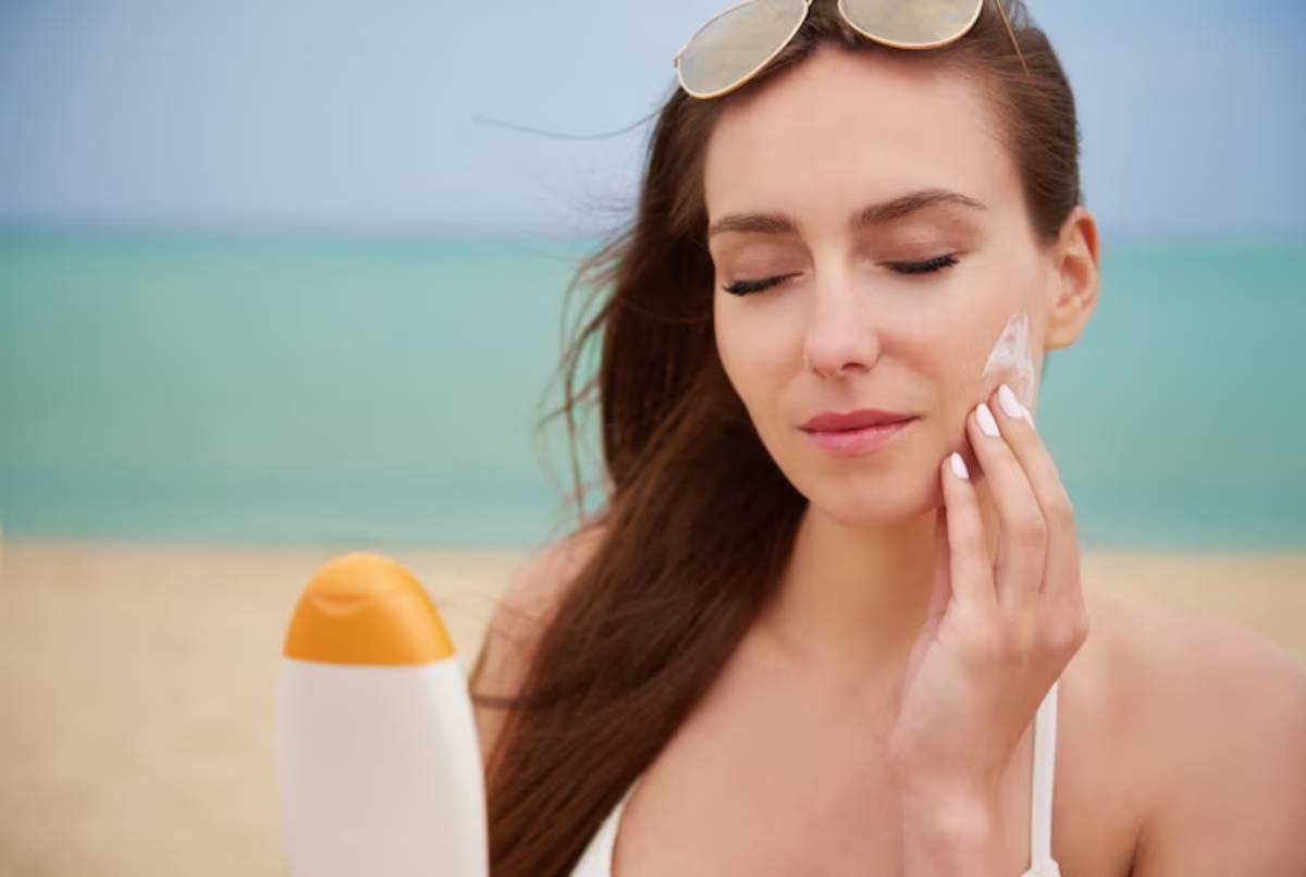 A young woman in a white swimsuit holds a sunscreen bottle at the beach, with the ocean and sky in the background.