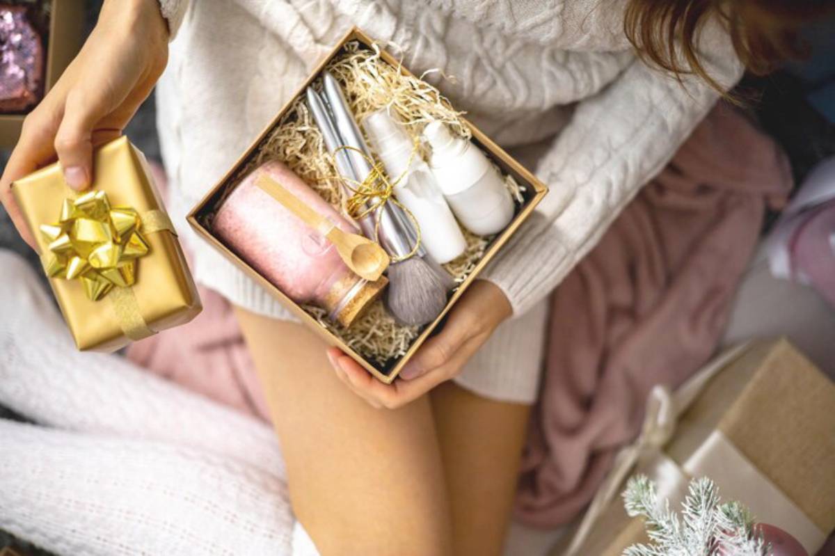 A person in a cozy sweater holds a gift box containing beauty products, with a gold-wrapped present in their other hand.