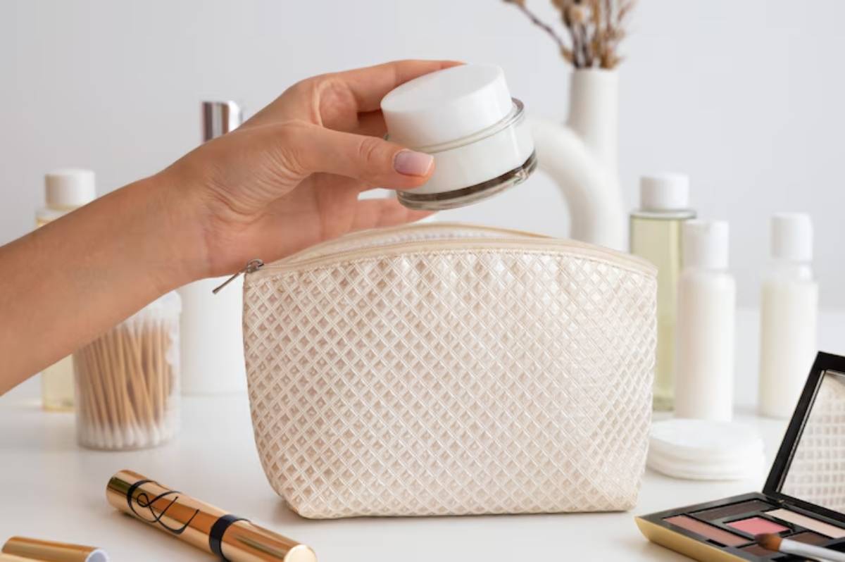 A hand holds a cream jar above a textured beige cosmetic bag, surrounded by various skincare and makeup products on a light surface.
