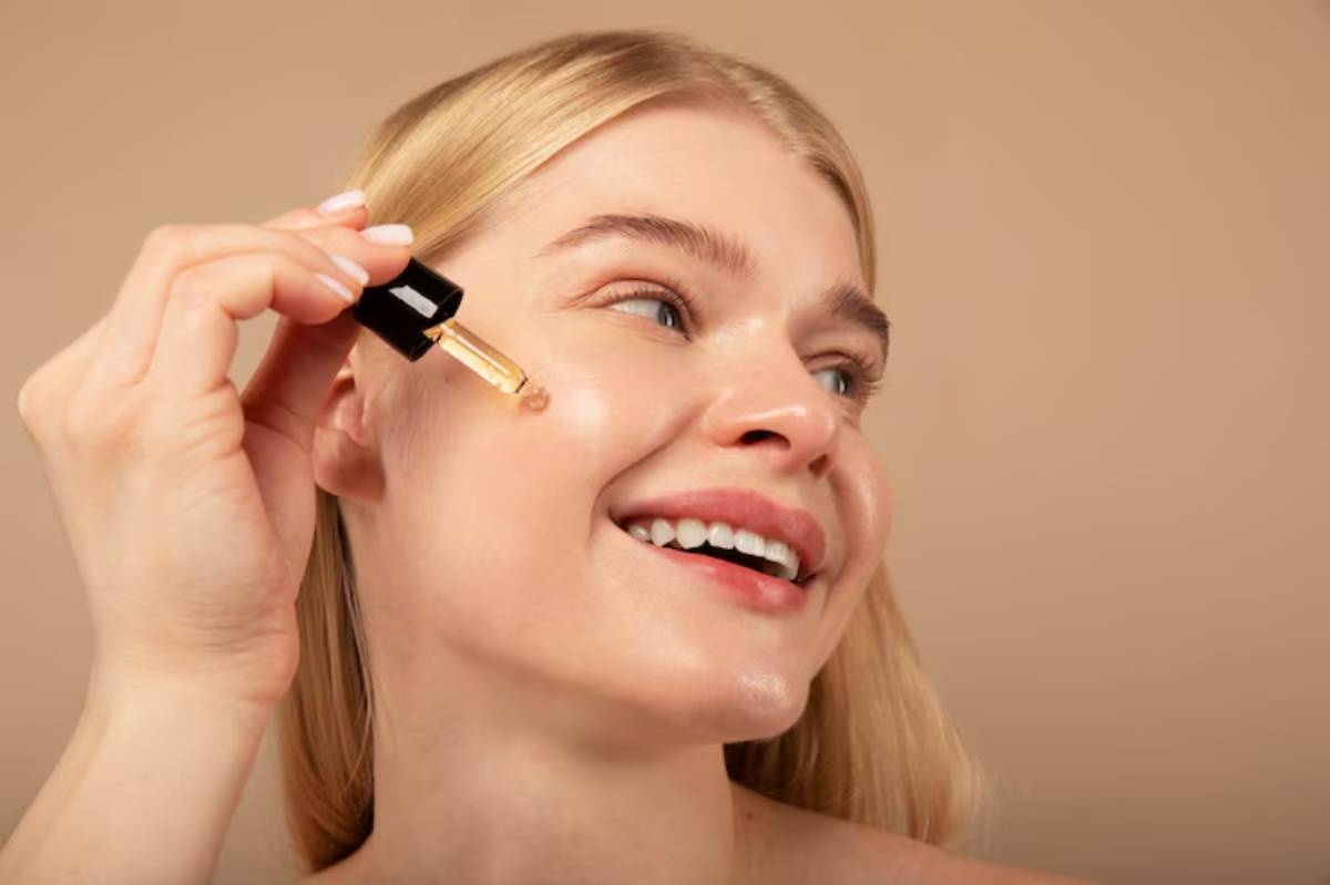 A woman with long blonde hair applies makeup, holding a cosmetic product with a neutral background.