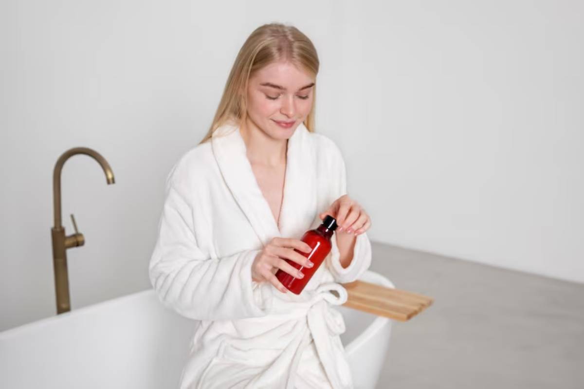 A person in a white bathrobe holds a red bottle while sitting beside a modern bathtub in a minimalist bathroom setting.