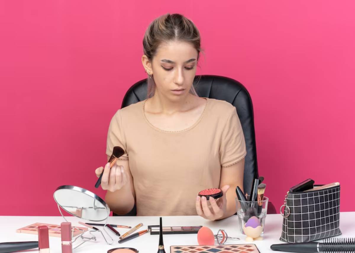 A person holding a makeup brush and a compact, surrounded by various beauty products on a table with a pink background.