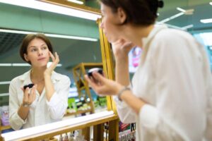 A woman in a white shirt applies makeup while looking into a mirror in a bright, vibrant cosmetics store.