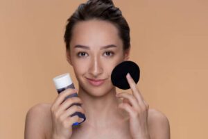 A woman holds a blue bottle of skincare product in one hand and a black cotton pad in the other, against a peach background.