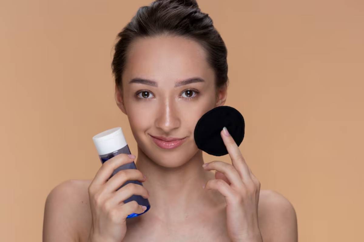 A woman holds a blue bottle of skincare product in one hand and a black cotton pad in the other, against a peach background.