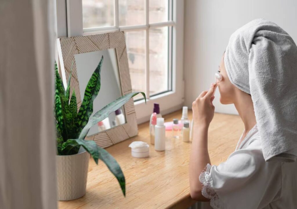 A woman in a white robe and towel applies skincare in front of a mirror, surrounded by beauty products and a potted plant.