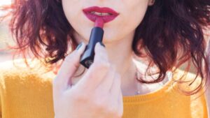 A woman with curly reddish hair applies deep red lipstick while wearing a bright yellow sweater, focusing on her lips.