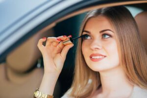 A woman holding a lipstick tube while sitting in a car, with well-manicured red nails and flowing, wavy hair.