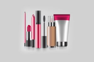 A lineup of various makeup products including lipsticks, a lip pencil, mascara, foundation, and cream, displayed on a light gray background.