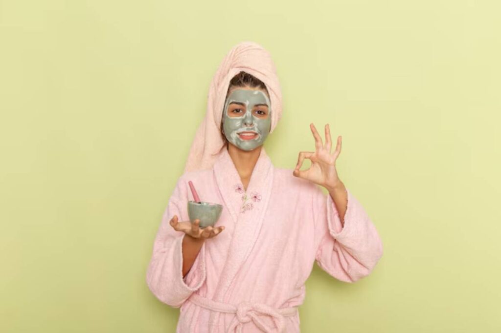 A person in a pink bathrobe and towel holds a small bowl in one hand and makes an okay gesture with the other against a green wall.
