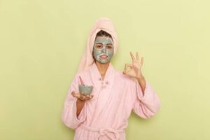 A person in a pink bathrobe and towel holds a small bowl in one hand and makes an okay gesture with the other against a green wall.