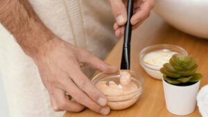 A hand with a brush applies peach-colored cream from a bowl on a wooden countertop, with a small plant and another cream jar nearby.
