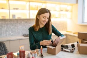 A woman in a green polka dot blouse organizes makeup products inside a cardboard box on a marble countertop.