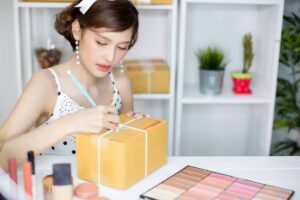 A person in a polka dot dress writes on a wrapped package, surrounded by makeup products and plants on a bright workspace.