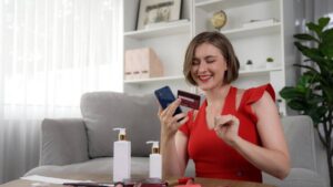 A woman in a red dress sits on a couch, holding a phone and a credit card, surrounded by beauty products on a table.